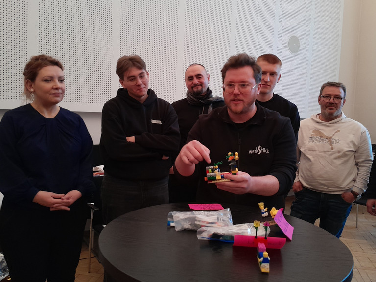 Ein Mann spricht zur umstehenden Gruppe an einem Tisch mit einem Segelboot aus bunten Legosteinen beim Teamtag vom Meisterbetrieb für Handwerk Werkstück Berlin zum Thema "Gute Kommunikation verbindet" in Berlin-Wannsee Ein Mann spricht zur umstehenden Gruppe an einem Tisch mit einem Segelboot aus bunten Legosteinen beim Teamtag vom Meisterbetrieb für Handwerk Werkstück Berlin zum Thema "Gute Kommunikation verbindet" in Berlin-Wannsee